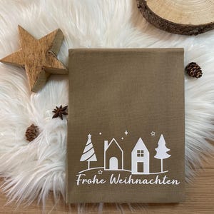 Könnte beinhalten: Eine braune Geschenkbox mit einer weißen Grafik von Häusern und Bäumen und dem Text "Frohe Weihnachten". Ein Holzstern, Tannenzapfen und eine Holzscheibe sind auf einer weißen, fellartigen Oberfläche angeordnet.