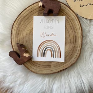 Puede incluir: Una figurita de madera de un oso y una ardilla sobre una rodaja de madera con una tarjeta que dice "Willkommen Kleines Wunder" y una ilustración de arcoíris.