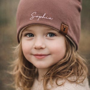 Könnte beinhalten: Eine altrosa Beanie-Mütze mit dem Namen Sophia in weißer Schreibschrift auf der Vorderseite. Die Mütze hat ein kleines braunes Kunstleder-Etikett an der Seitenkante, das mit einer schwarzen Silhouette eines Hirschkopfes geprägt ist.