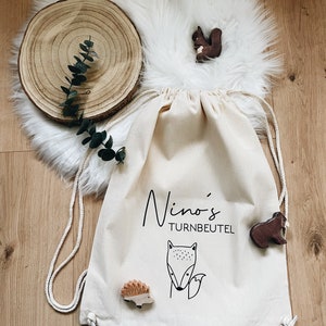 Puede incluir: Una bolsa de cord&oacute;n blanco con el texto "Nino's Turnbeutel" impreso en ella. La bolsa est&aacute; decorada con una ilustraci&oacute;n de zorro y erizo. Hay figuritas de animales de madera en la bolsa, incluyendo una ardilla, un erizo y un zorro.