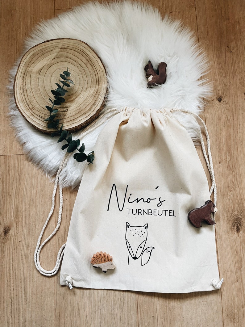 Puede incluir: Una bolsa de cord&oacute;n blanco con el texto "Nino's Turnbeutel" impreso en ella. La bolsa est&aacute; decorada con una ilustraci&oacute;n de zorro y erizo. Hay figuritas de animales de madera en la bolsa, incluyendo una ardilla, un erizo y un zorro.