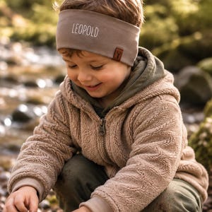 Puede incluir: Una cinta para la cabeza de color topo con el nombre LEOPOLD impreso en letras blancas en la parte frontal, con una pequeña etiqueta rectangular marrón en el lateral que muestra una silueta oscura de una cabeza de ciervo.
