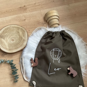 Puede incluir: Una bolsa de cord&oacute;n verde con un dise&ntilde;o de le&oacute;n blanco y el nombre "Leo" escrito debajo. La bolsa est&aacute; sobre una superficie blanca y esponjosa con una figurita de ardilla y oso de madera.