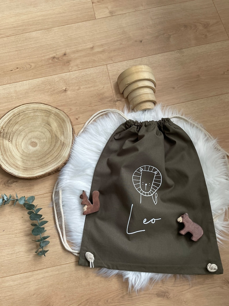 Puede incluir: Una bolsa de cord&oacute;n verde con un dise&ntilde;o de le&oacute;n blanco y el nombre "Leo" escrito debajo. La bolsa est&aacute; sobre una superficie blanca y esponjosa con una figurita de ardilla y oso de madera.