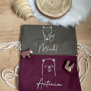 Puede incluir: Dos bolsas de cord&oacute;n personalizadas con ilustraciones de animales. La primera bolsa es de color verde oscuro con un zorro y el nombre "Mich" impreso en blanco. La segunda bolsa es de color burdeos con un oso y el nombre "Antonia" impreso en blanco.