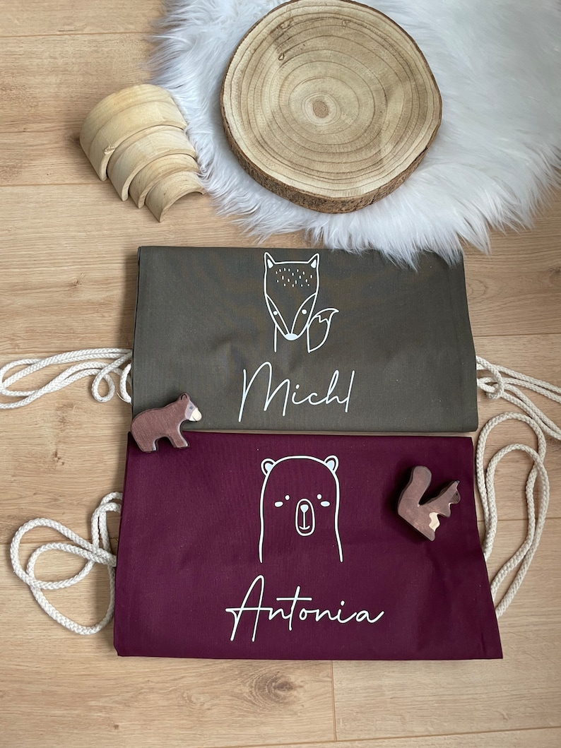 Puede incluir: Dos bolsas de cord&oacute;n personalizadas con ilustraciones de animales. La primera bolsa es de color verde oscuro con un zorro y el nombre "Mich" impreso en blanco. La segunda bolsa es de color burdeos con un oso y el nombre "Antonia" impreso en blanco.