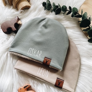 Puede incluir: Dos gorros suaves, uno verde salvia y otro beige, cada uno con un nombre impreso en blanco, 'OSKAR' en el verde y 'LEON' en el beige, con una pequeña etiqueta de cuero sintético marrón que muestra un icono de silueta de cabeza de ciervo.
