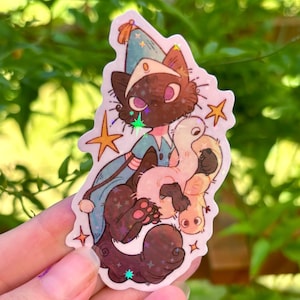 May include: A holographic sticker featuring a dark brown cat wearing a blue pointed hat and cape, holding a pale pink worm-like creature. The design includes orange stars and a star-shaped holographic effect across the surface of the sticker.
