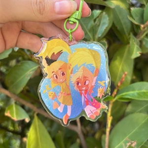Puede incluir: Un llavero acrílico transparente con dos personajes de dibujos animados con pelo y atuendos amarillos. Los personajes están sobre un fondo azul con detalles amarillos. El llavero tiene un cierre verde.