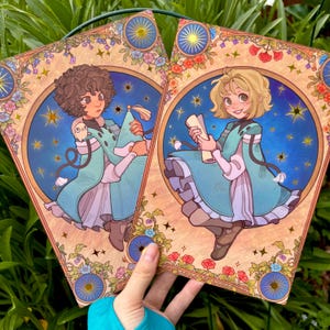 May include: Two art prints featuring illustrations of figures in teal robes, holding objects. The figures are set against a starry blue background within a decorative frame with floral and celestial elements. The prints are held up by a hand.