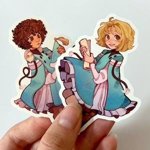 May include: Two die-cut stickers featuring illustrations of characters in teal and pink robes. One character has brown curly hair and the other has blonde hair. Both are holding objects and have black accents.