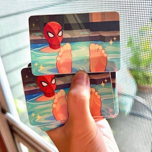 May include: A rectangular sticker featuring a cartoon illustration of a red-masked superhero in a swimming pool with two feet visible in the foreground. The water is light blue with sparkling yellow star accents and a dark tiled pool edge.
