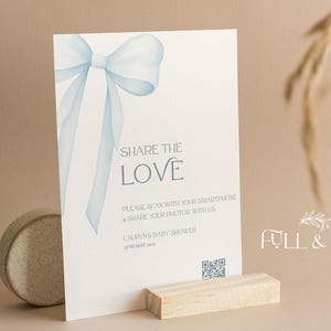 May include: A baby shower invitation card with a light blue bow illustration and the text "SHARE THE LOVE". The card is displayed on a wooden stand. The words "FULL & LOVE" are also visible.
