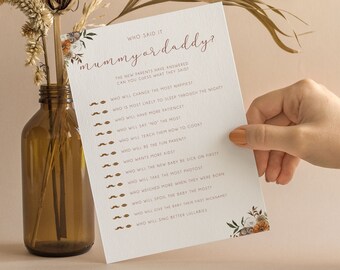May include: A white rectangular baby shower game card titled Who Said It Mummy or Daddy? with brown text and mustache icons. The card features floral illustrations in the corners and lists questions about parenting. The paper has a textured finish.