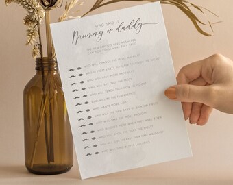 May include: A white card with the text "Who said it? Mommy or Daddy" in black script. The card has a list of questions about new parents and their baby, such as "Who will change the most diapers?" and "Who will be the sun parent?" The card is held in a hand against a light brown background.