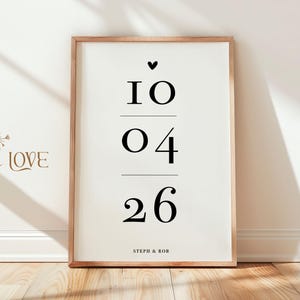 May include: A vertical rectangular framed print with a white background and black text. The design features a small heart icon above the date 10 04 26, separated by horizontal lines. The names STEPH & ROB are printed in small black letters at the bottom.