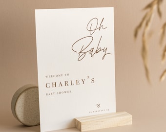May include: A rectangular white sign with brown text that reads Oh Baby, Welcome to Charley's Baby Shower, and 12 February 23, featuring a small heart icon. The sign is supported by a rectangular light wood block stand.