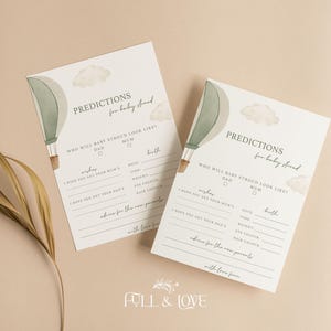 May include: Two baby shower prediction cards with a hot air balloon and cloud design. The cards have prompts for guests to fill out, including predictions for the baby's appearance and advice for the parents. The cards are on a neutral background.