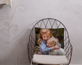 May include: Two young girls are hugging each other while smiling. They are sitting on a chair with a white cushion. The chair has a black metal frame.