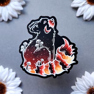 May include: A die-cut sticker featuring a black bear silhouette with a starry night sky and mushroom design. The bear is surrounded by flames in shades of red, orange, and yellow. The sticker is on a gray background with white flowers.