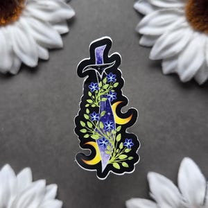 May include: A sticker featuring a stylized sword design with a purple blade and black hilt. The sword is adorned with blue flowers, green leaves, and yellow crescent moons. The sticker is surrounded by white sunflowers on a gray background.