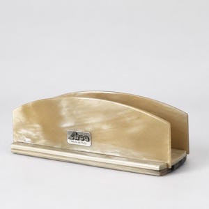 Puede incluir: Un servilletero rectangular con borde superior curvado, fabricado en un material jaspeado de color beige y crema. En la parte frontal, centrada, hay una pequeña placa metálica rectangular de color plateado con el texto "Area" y "Made in Italy" en negro.