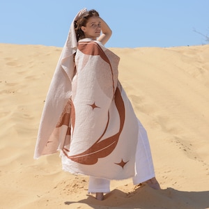 May include: A large, rectangular, textured cotton throw blanket featuring a cream-colored base with a prominent rust-colored graphic print of a crescent moon, sun rays, and four-pointed stars. The blanket has a frayed edge finish.