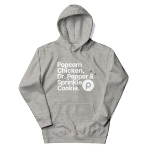Puede incluir: Una sudadera con capucha gris con un bolsillo delantero y un cordón. La parte delantera de la sudadera presenta el texto "Popcorn Chicken, Dr. Pepper & Sprinkle Cookie." y un logotipo "P" blanco.