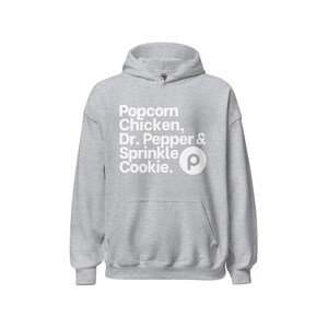 Puede incluir: Una sudadera con capucha gris claro con un bolsillo delantero y un cordón. La parte delantera de la sudadera presenta las palabras "Popcorn, Chicken, Dr. Pepper & Sprinkle Cookie". También es visible un logotipo "P" blanco.
