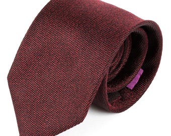 May include: A burgundy tie with a textured weave, rolled up to show its shape. The tie has a deep red color with subtle dark accents. A small purple tag is visible on the back of the tie.