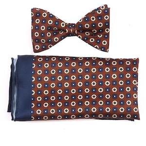 May include: A brown and blue patterned bow tie and matching pocket square. The bow tie and pocket square have a repeating pattern of small squares with a white centre.