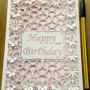 May include: A white card with a pink and white intricate design. The card has a white cutout with the text "Happy Birthday" in pink.