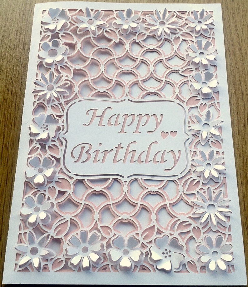 May include: White card with a pink and white intricate cut-out design. The card says "Happy Birthday" in a white script font.