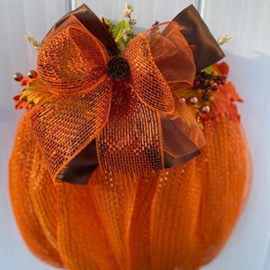 May include: A pumpkin-shaped wreath made of orange mesh material, featuring a large decorative bow crafted from layered orange mesh and brown satin ribbons. A small pinecone sits at the center of the bow, surrounded by artificial autumn leaves and berries.