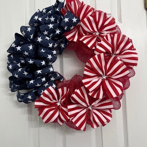 May include: A red, white, and blue patriotic wreath with a star-spangled banner design. The wreath is made of mesh and ribbon and features red, white, and blue bows.