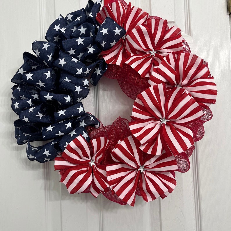 May include: A red, white, and blue patriotic wreath with a star-spangled banner design. The wreath is made of mesh and ribbon and features red, white, and blue bows.