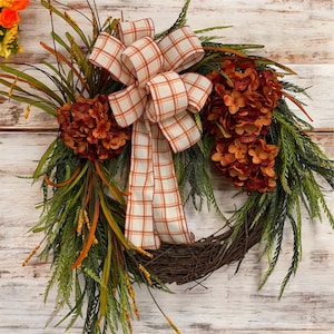 May include: A fall-themed wreath with a brown grapevine base, adorned with orange and red hydrangeas, green foliage, and a plaid bow with orange and white squares. The wreath is a decorative piece for the home.