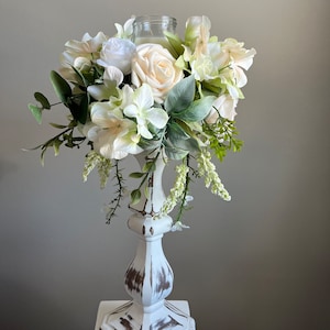 May include: White candle holder with a distressed white finish. The holder is decorated with a ring of white flowers and greenery.