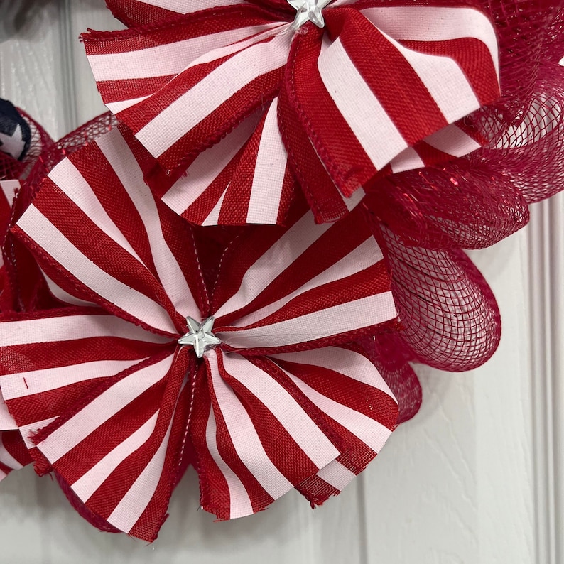 May include: A red and white striped fabric bow with a silver star in the center. The bow is attached to a red mesh wreath.