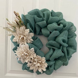 May include: A teal burlap wreath with three off-white fabric flowers and dried grasses.