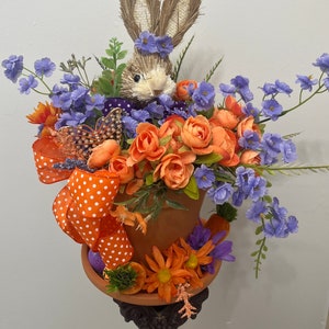 May include: A terracotta flower pot with a bunny rabbit figurine, orange and purple flowers, and a white polka dot orange ribbon. The pot is sitting on a black pedestal.