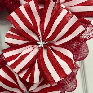 May include: A red and white striped ribbon bow with a silver star in the center. The bow is part of a wreath made with red mesh.