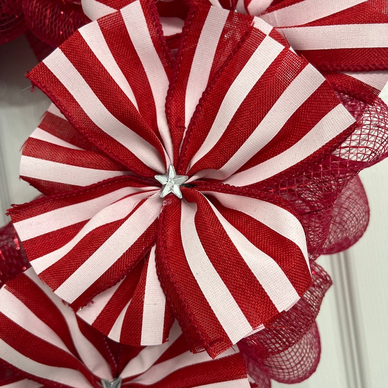 May include: A red and white striped ribbon bow with a silver star in the center. The bow is part of a wreath made with red mesh.