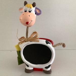 May include: A white ceramic cow planter with black spots. The cow has a red base and a chalkboard on its back. The cow's head is separate and has a yellow and purple design. The cow is holding a piece of chalk and a small green bag.