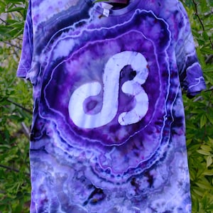 Op de afbeelding: Een paars tie-dye t-shirt met een groot wit "dB" logo. Het shirt hangt aan een witte hanger tegen een achtergrond van groen gebladerte. Het label luidt "IN THERE ART".