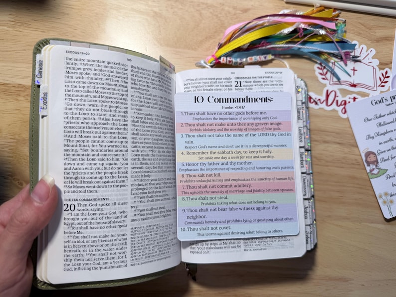 Może przedstawiać: Otwarta Biblia z zieloną okładką, wyświetlająca tekst z Księgi Wyjścia i zakładkę z dziesięcioma przykazaniami. Widoczne są kolorowe wstążki i ozdobna naklejka.