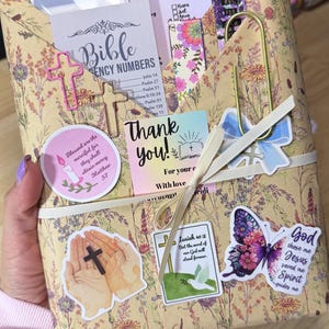 May include: A tan envelope with a floral print containing religious cards and stickers. It features text including Books of the Bible, Bible Emergency Numbers, Thank you, and Isaiah 40:8. A gold paperclip and a white ribbon are attached to the front.