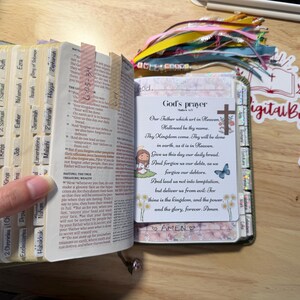 Może przedstawiać: Otwarta Biblia z oznaczonymi zakładkami i ozdobną zakładką. Strony są oznaczone kolorowymi zakładkami. Widoczna jest karta z tekstem "God's prayer" i ilustracją krzyża.