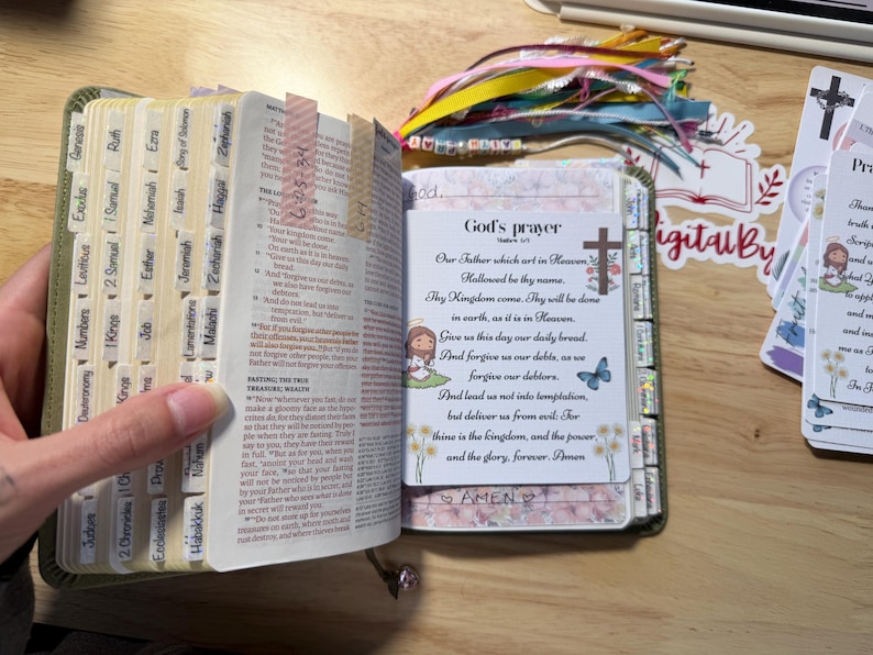 Może przedstawiać: Otwarta Biblia z oznaczonymi zakładkami i ozdobną zakładką. Strony są oznaczone kolorowymi zakładkami. Widoczna jest karta z tekstem "God's prayer" i ilustracją krzyża.