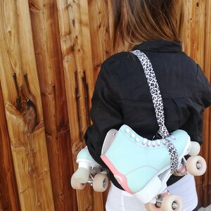 May include: A person wearing a black shirt and carrying a pair of light blue roller skates with white wheels and pink accents. The skates are attached to a leopard print strap.